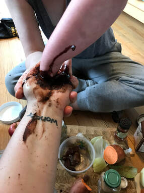 Onions, cinnamon, apples, and garlic lay on a wooden cutting board. Above, Alexis' stump is covered in chocolate sauce that she mixed in Lindsay's upturned hand. Lindsay's wrist tattoo is visible, and it reads 