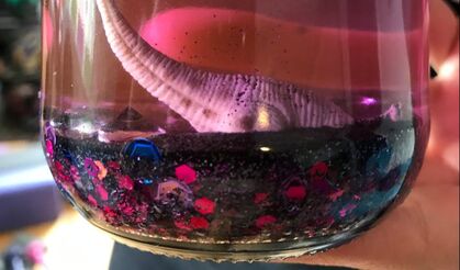 A hand holds a sensory jar, filled with pink liquid. The bottom of the jar is layered with sparkles. A toy dinosaur buries its head in the sparkly sand